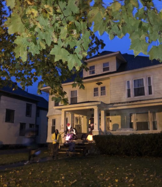 Visual: Uma casa de madeira de dois anderas e sotão, moldada pela folhagem da arvore de mapple no jardim. Não tem muros ou grades. A casa fica no topo de um gramado. É um final de tarde de céu azul. 